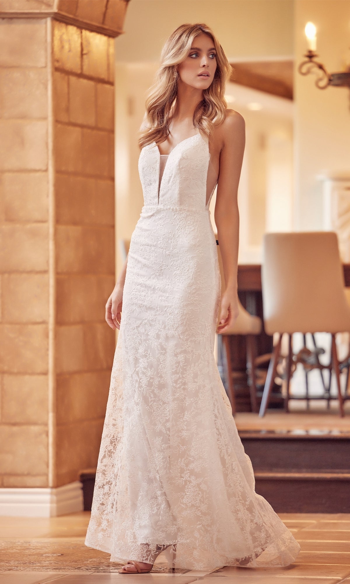 Wearing the Backless Long Off-White Lace Formal Dress 272W, a woman stands indoors near a stone wall and barstools, looking to the side.